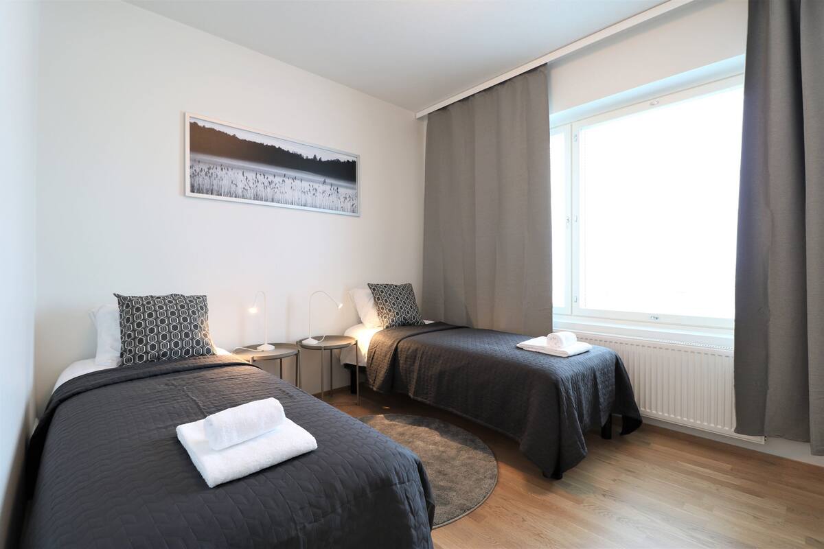 A hotel room with two beds, a round rug, a bedside table with a lamp, a window with curtains, and a wall-mounted picture.