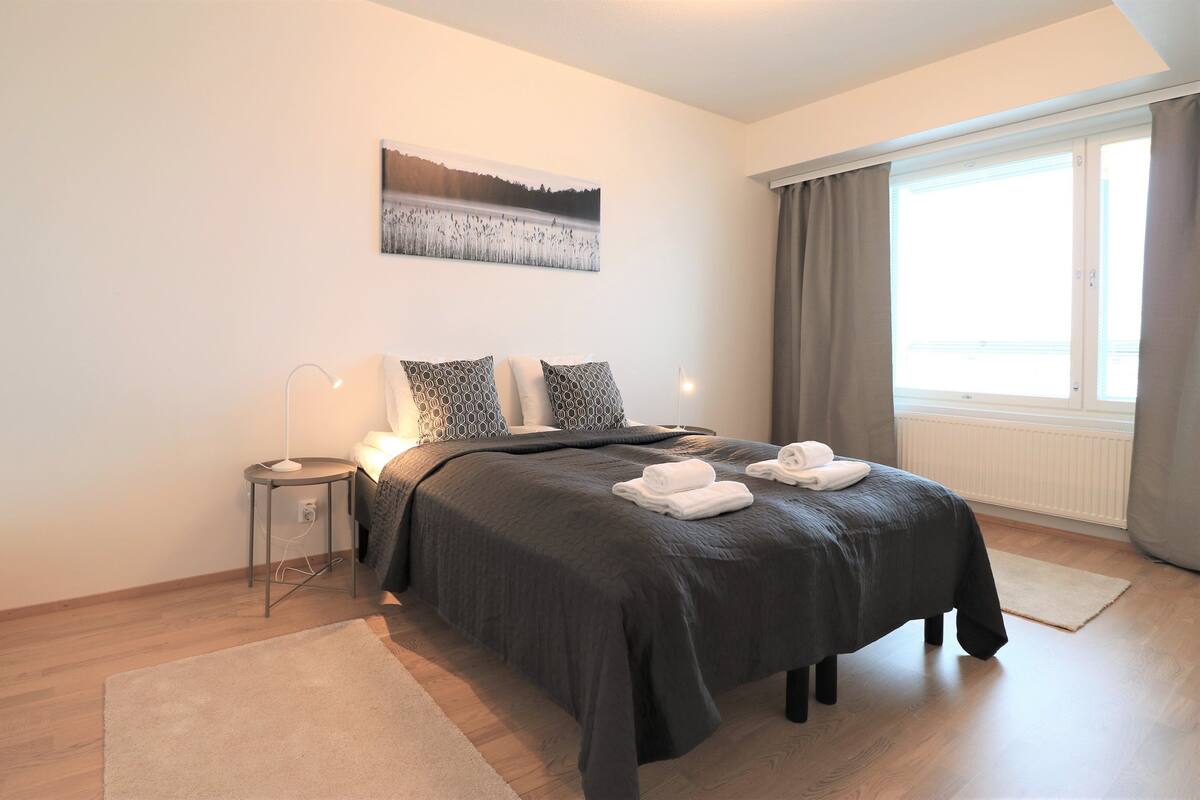 A modern bedroom with a large bed, a bedside table with a lamp, a painting on the wall, and a window with curtains.