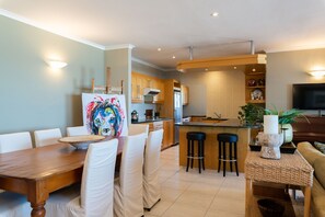 A modern kitchen with wooden cabinets, a dining area with a wooden table and white chairs, and a small bar area with stools. - Lagoon Beach Penthouse (Cape Town)