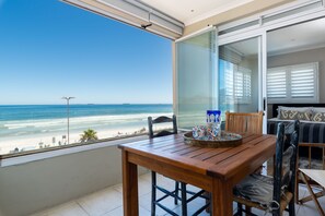 A balcony with a wooden table and chairs, overlooking a beach with a clear view of the ocean and distant shoreline. - Lagoon Beach Penthouse (Cape Town)