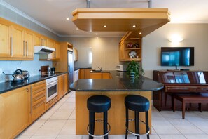 A modern kitchen with wooden cabinets, a central island with stools, a built-in oven, and a television on the wall. - Lagoon Beach Penthouse (Cape Town)