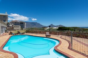 A swimming pool with a curved edge, surrounded by a brick patio and a metal fence. - Lagoon Beach Penthouse (Cape Town)