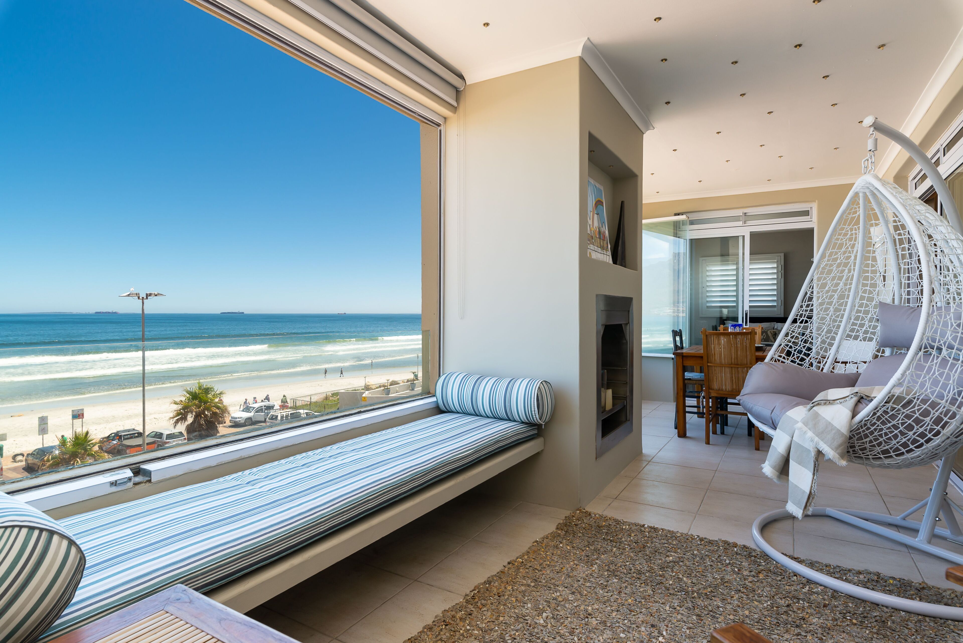 A room with a large window overlooking a beach, a hanging chair, a sofa, and a small table.