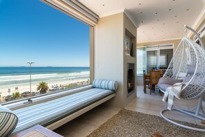 Quarto com uma grande janela com vista para a praia, uma cadeira suspensa, um sofá e uma pequena mesa.