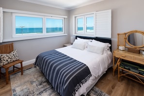 Quarto com uma cama grande, duas janelas com vista para o oceano, uma mesinha de cabeceira de madeira, uma cadeira de vime e uma mesa de apoio com espelho.