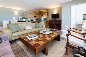 A modern living room with a wooden coffee table, a beige sofa with patterned cushions, a wooden dining table, and a kitchen area in the background. - Lagoon Beach Penthouse (Cape Town)