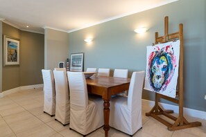 A dining area with a wooden table surrounded by chairs, a painting on easel, and a framed picture on the wall. - Lagoon Beach Penthouse (Cape Town)