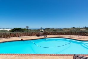 A clear blue swimming pool with a wooden deck and a bench, surrounded by a brick border and a metal fence. - Lagoon Beach Penthouse (Cape Town)