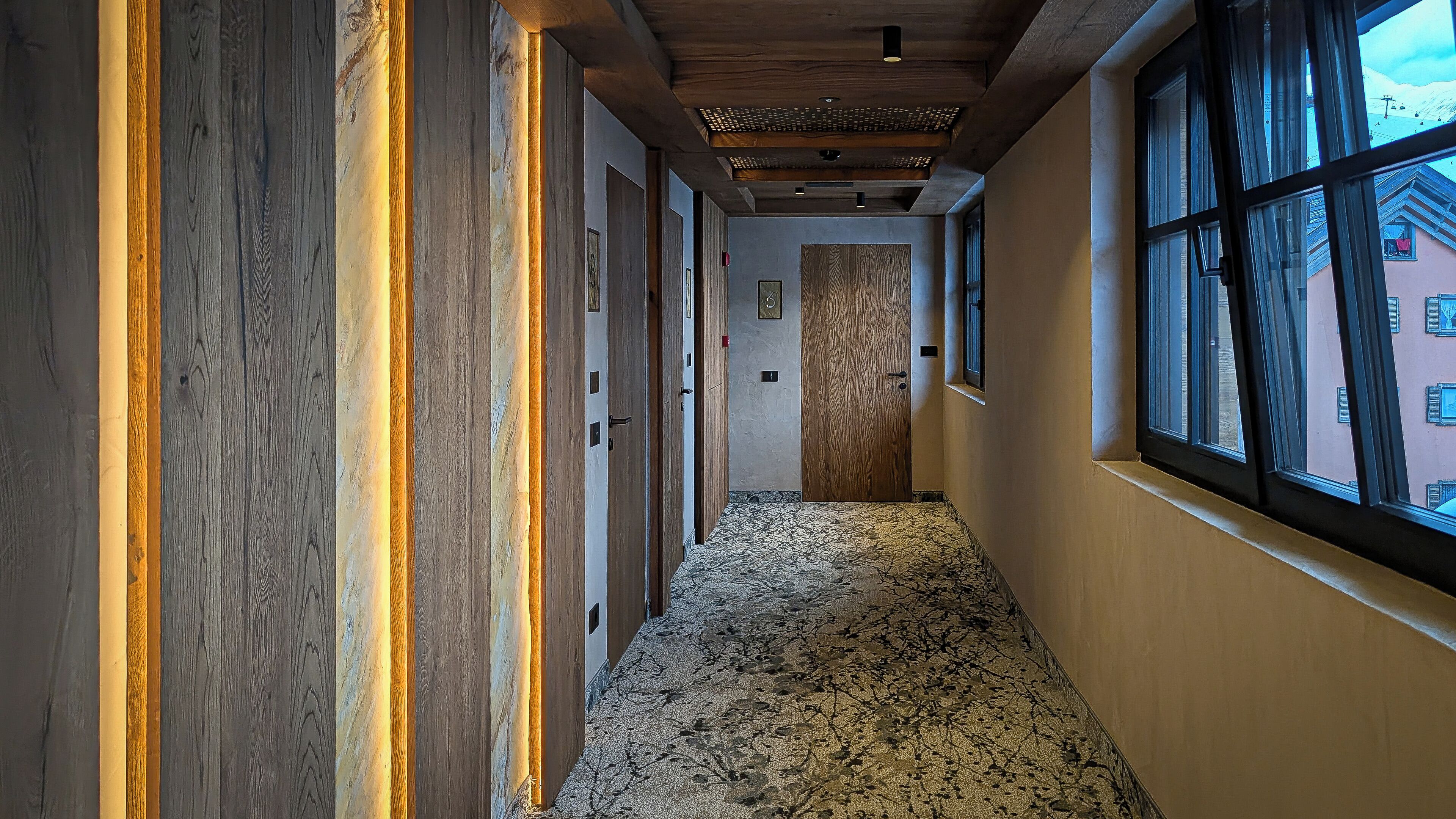A hallway with wooden paneling, a patterned carpet, and large windows with a view of buildings outside.