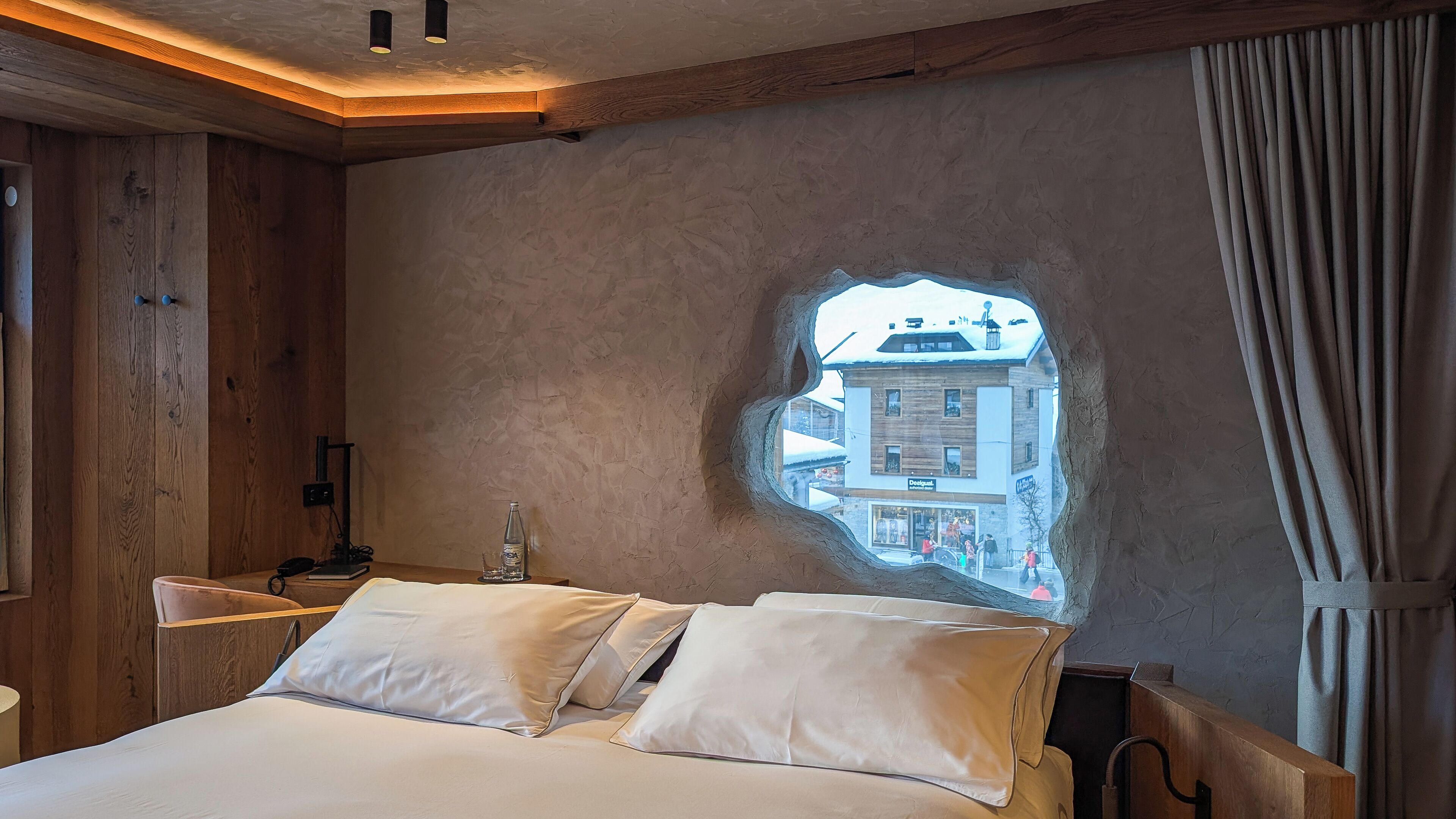 A bedroom with a large bed, a wooden headboard, a nightstand, and a window with a view of buildings outside.