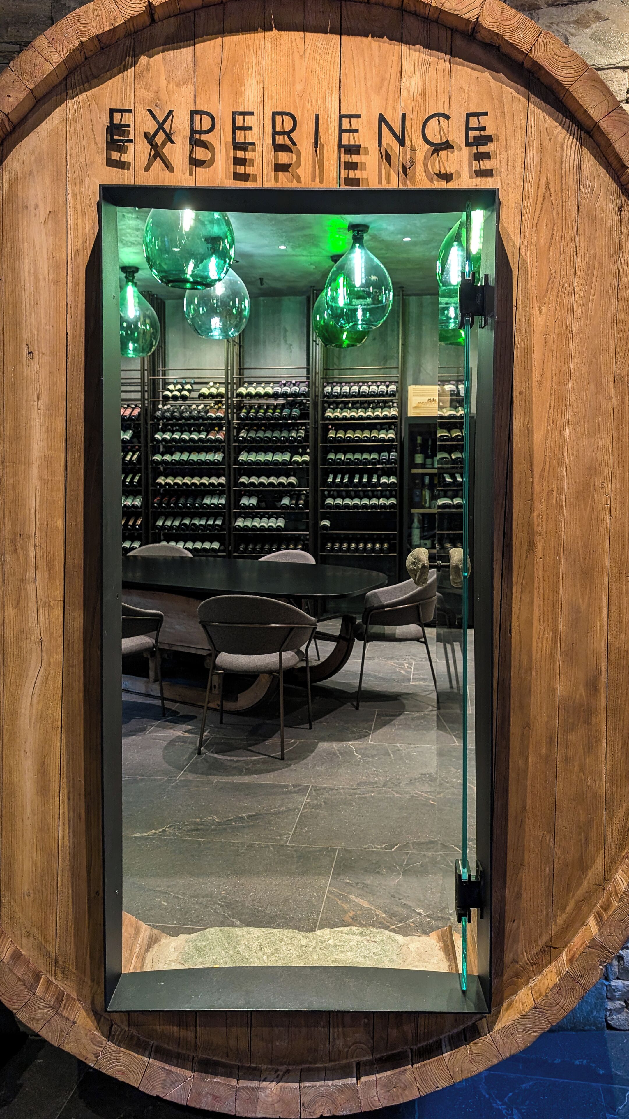 A wooden barrel-shaped entrance with the word "EXPERIENCE" above it. Inside, there are shelves with wine bottles, a table, chairs, and hanging green lights.