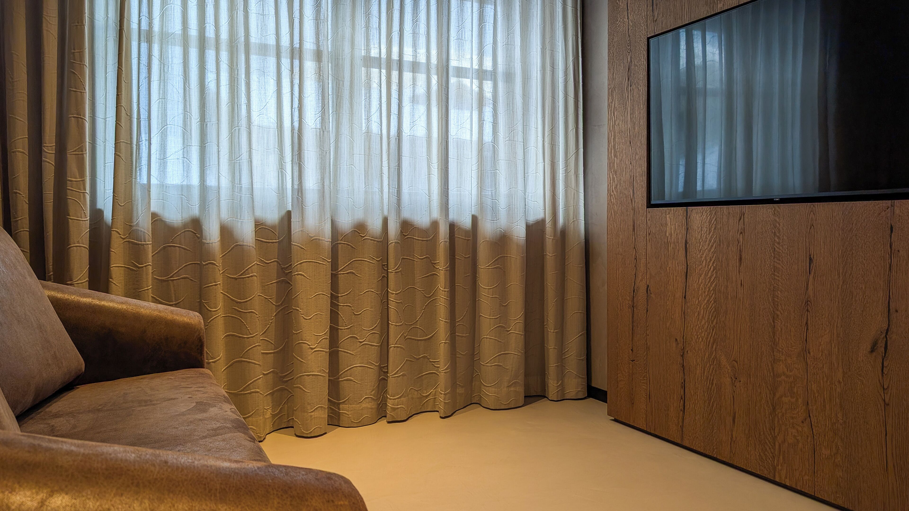 Una sala de estar moderna con un sofá, un televisor instalado en una pared de madera y cortinas de gasa.