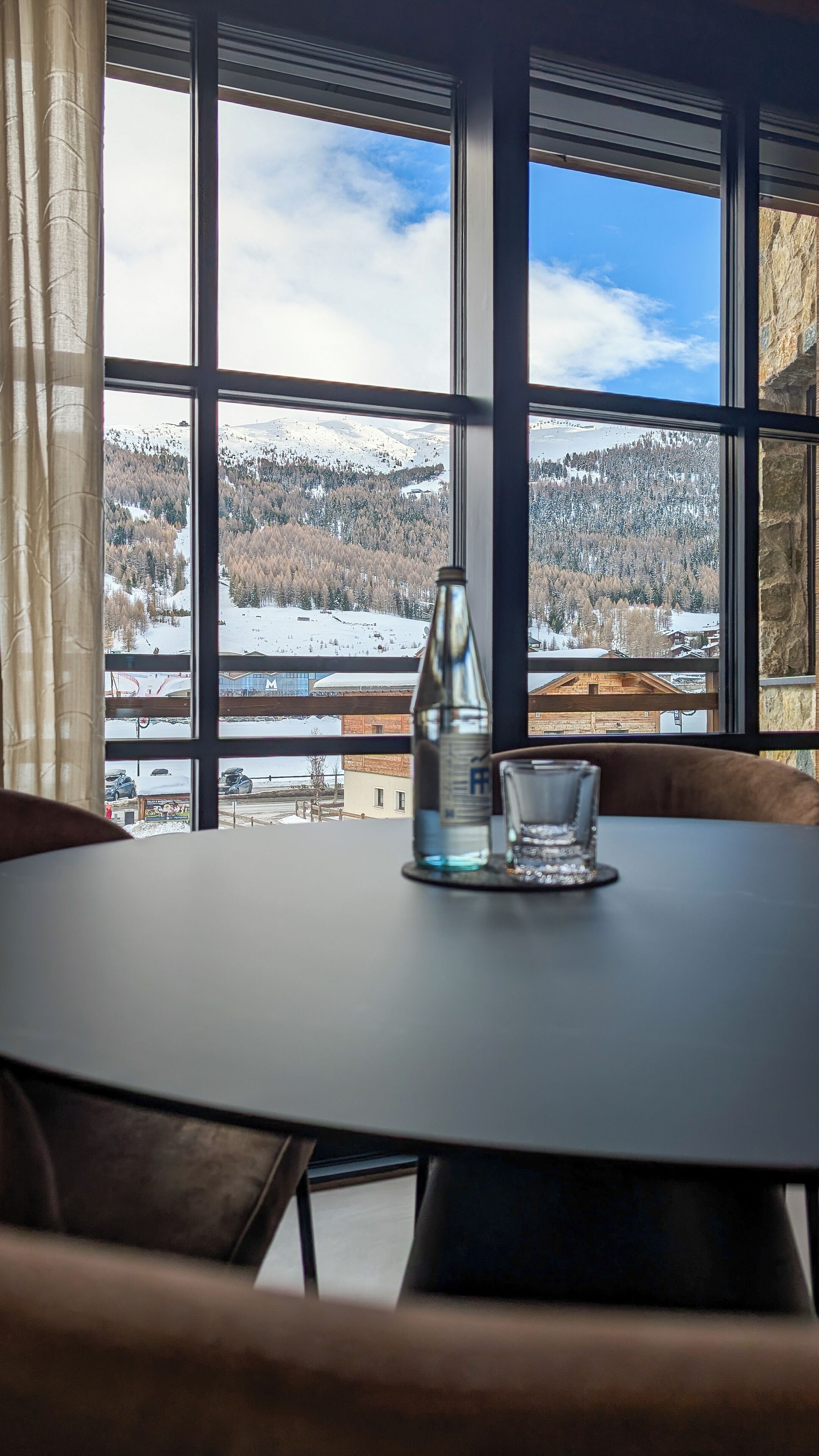 Une table avec une bouteille et un verre, une fenêtre donnant sur un paysage enneigé et un rideau.