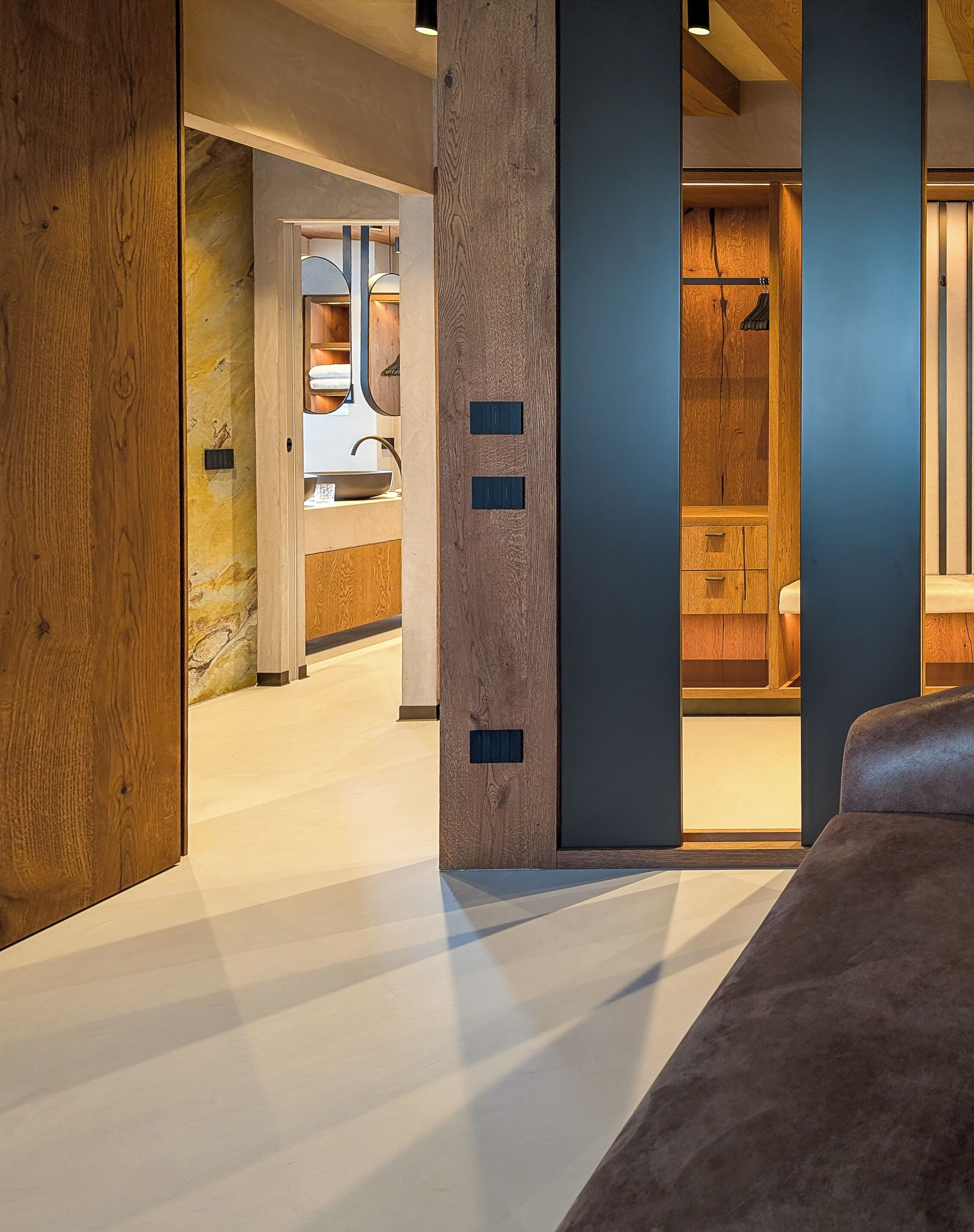 A modern interior with a wooden floor, a large central column, and a view into a room with a sink and cabinets.