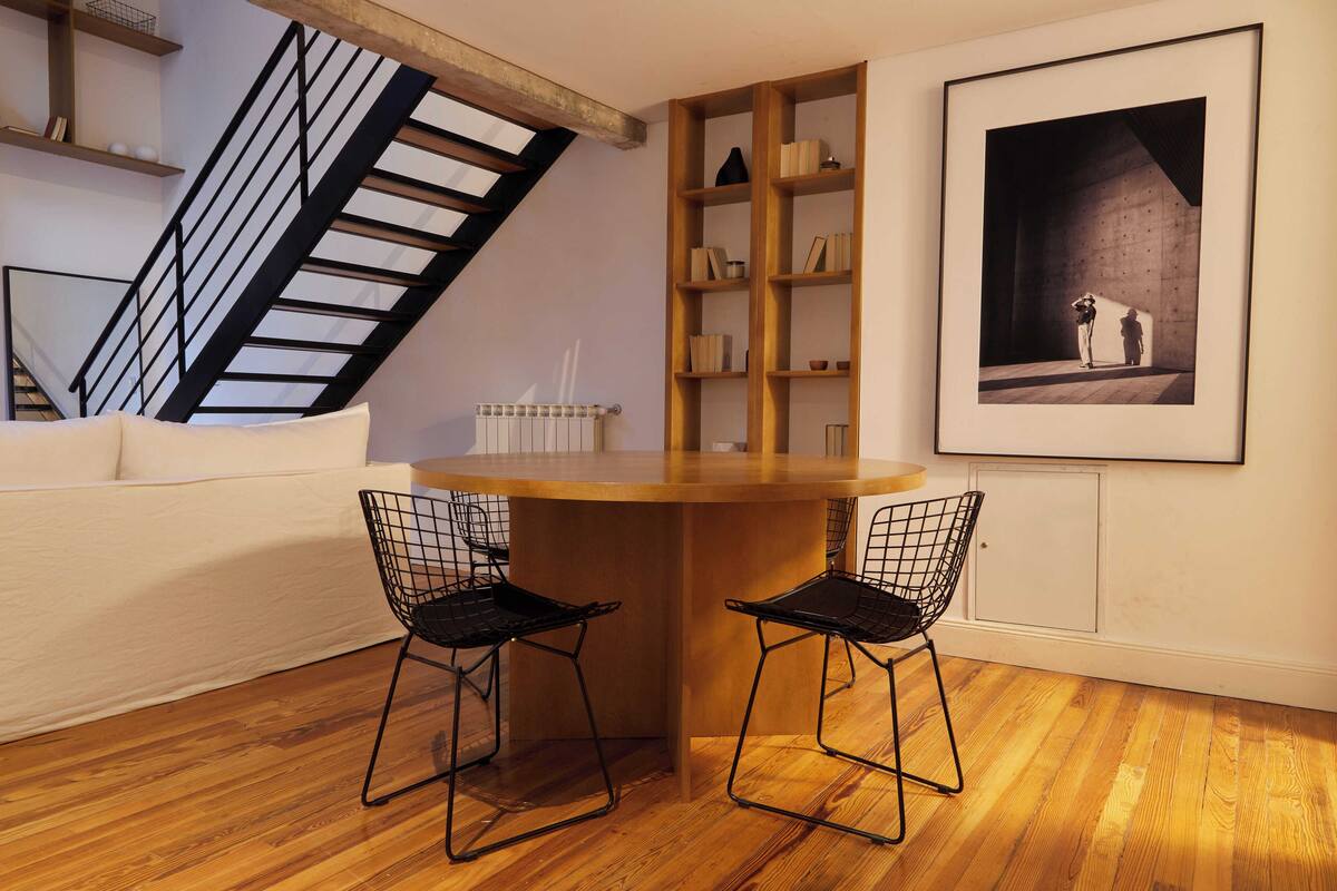 A modern living room with a wooden dining table, two black wire chairs, a white sofa, a wooden bookshelf, a framed artwork, and a staircase with a black railing.