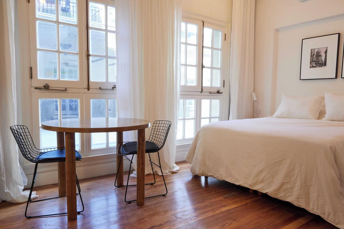 A hotel room with a large bed, a round wooden table with two chairs, and multiple windows with curtains.