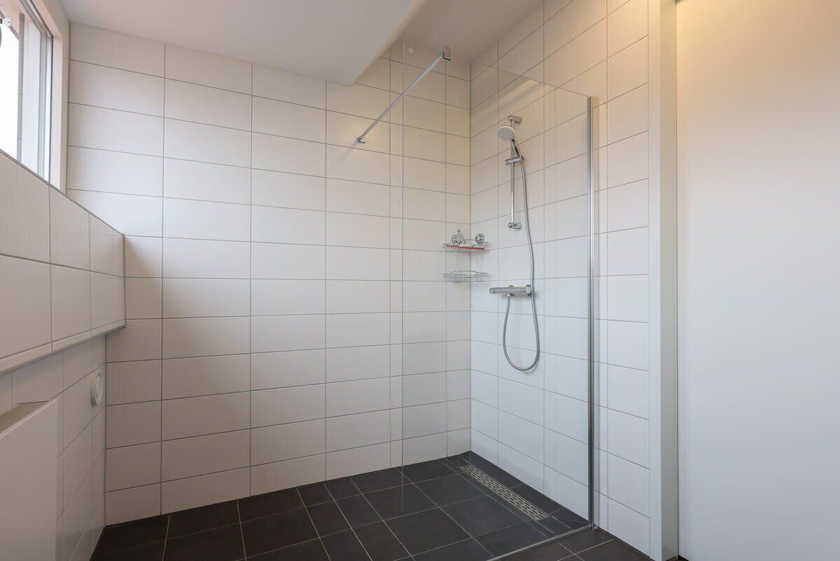 A bathroom with white tiled walls, a black tiled floor, a shower with a handheld showerhead, and a glass shower door.