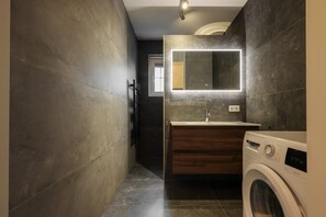 A modern bathroom with dark tiled walls, a wooden vanity, a built-in washing machine, and a large mirror with integrated lighting.