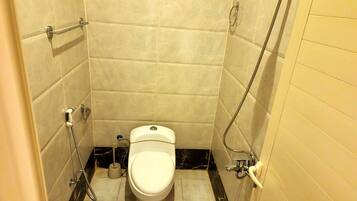 A small, compact bathroom with a toilet, a handheld showerhead, and a toilet brush.