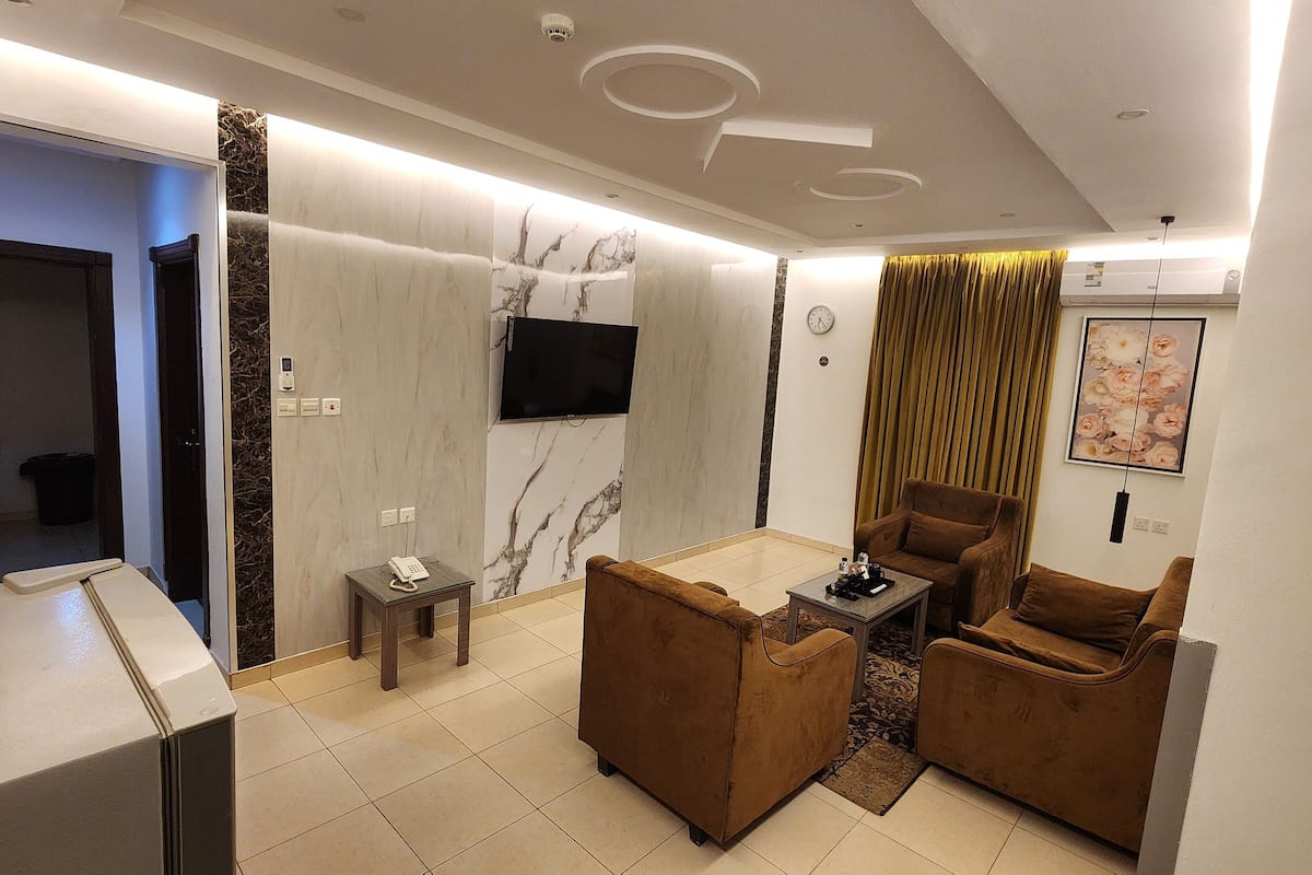 A modern living room with a sofa set, a coffee table, a flat-screen TV mounted on the wall, and a small side table with a phone.