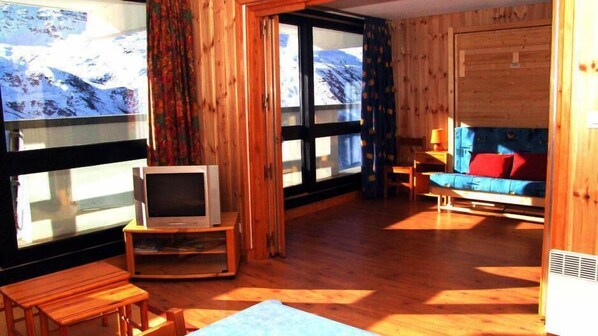 A wooden interior with a TV on a stand, a sofa with red cushions, and a window with a view of a snowy landscape.