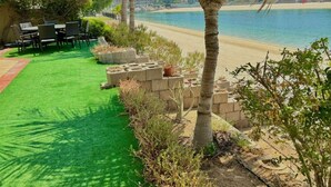 Zona junto a la piscina con césped artificial, asientos y vista a la playa.