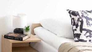Una cama bien tendida con sábana blanca y almohada de estampado blanco y negro. Una pequeña mesita de noche con una lámpara, un libro y una planta pequeña está al lado de la cama.
