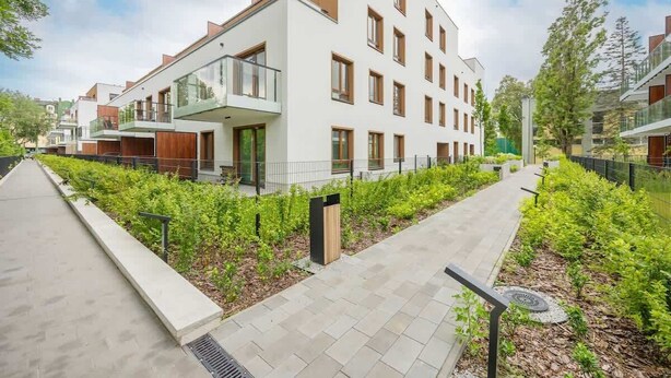 Un edificio residencial moderno de varios pisos con balcones, un paseo pavimentado y jardines.