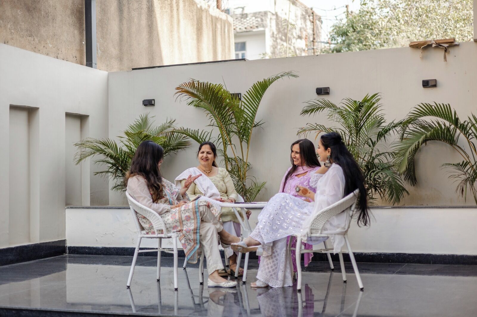 Un groupe de personnes est assis autour d’une table dans un espace extérieur aménagé en patio, avec des murs blancs et des plantes en pot.