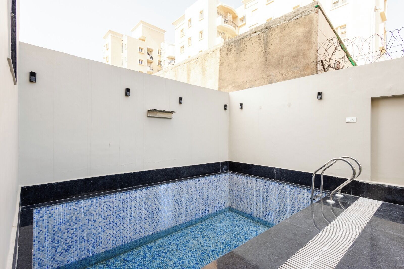 A small indoor pool with blue mosaic tiles, a metal ladder, and a white wall with a small shelf.