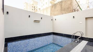 A small indoor pool with blue mosaic tiles, a metal ladder, and a white wall with a small shelf.