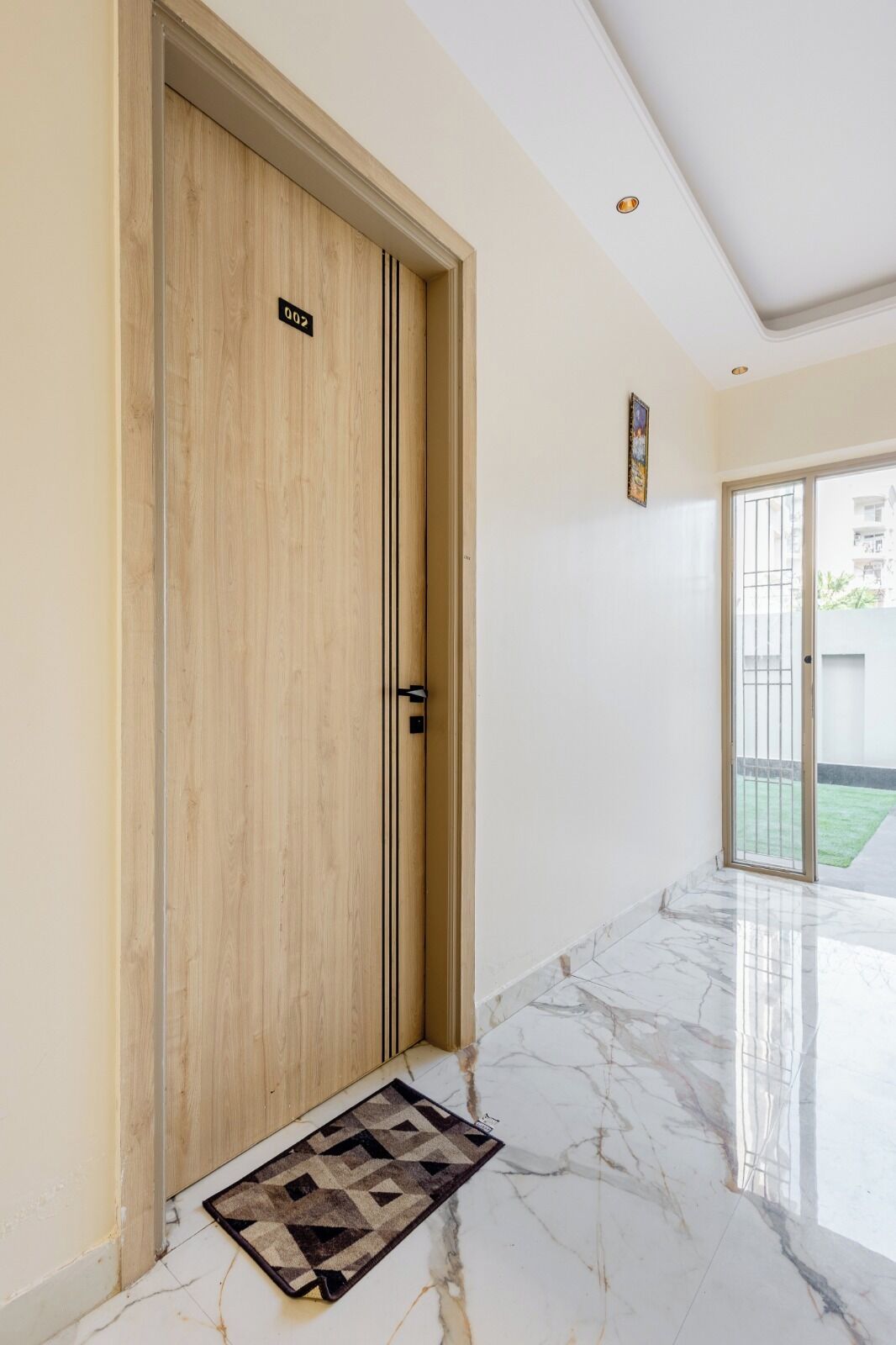 A hallway with a wooden door marked "002", a marble floor, and a view of a green outdoor area through a glass door.