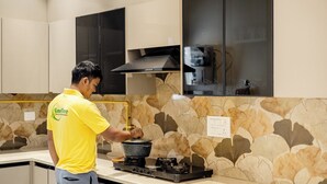 A person cooking on a stove in a modern kitchen with stone-patterned backsplash and light-colored cabinetry. - Lime Tree B&B Pari Chowk (Greater Noida)