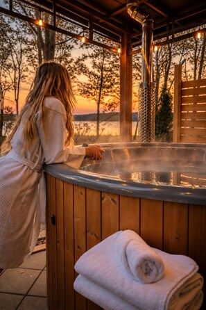 A person in a bathrobe leaning on a wooden spa tub with a towel beside it, set against a backdrop of trees and a sunset.