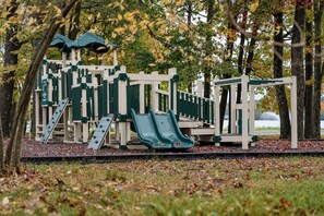 A playground with slides and climbing structures, surrounded by trees and a body of water in the background.