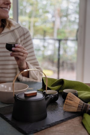 A person holding a cup, a tea kettle, and a whisk on a table with a green cloth.