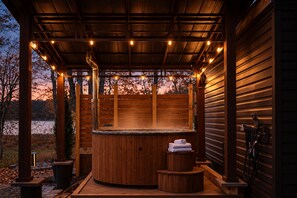A wooden outdoor hot tub area with a covered roof, surrounded by wooden walls, and a view of trees and a body of water in the background. - Autumn Lakefront: Natural Hot Tub: Private Dock: Play Area (Pocono Summit)