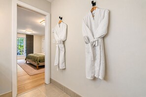 A hallway with two white robes hanging on hooks, a bed with a green blanket, and a view of a room with a window. - Autumn Lakefront: Natural Hot Tub: Private Dock: Play Area (Pocono Summit)