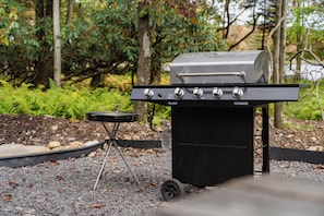 A portable barbecue grill with four burners and a side burner, placed on a gravel surface. - Autumn Lakefront: Natural Hot Tub: Private Dock: Play Area (Pocono Summit)