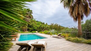 A wooden deck with a pool, lounge chairs, and palm trees. - VILLA NIDAZUR  VI4413 By Riviera Holiday Homes (Niza)