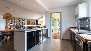 A modern kitchen with a central island, built-in appliances, and a dining area with chairs and a bookshelf. - VILLA NIDAZUR  VI4413 By Riviera Holiday Homes (Niza)