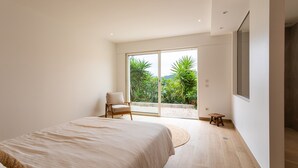 A modern bedroom with a large bed, a chair, a small stool, and a sliding glass door leading to a balcony with greenery. - VILLA NIDAZUR  VI4413 By Riviera Holiday Homes (Niza)