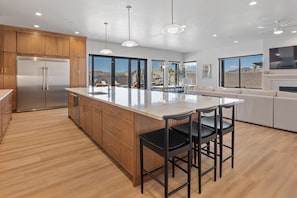 A modern kitchen with wooden cabinets, a stainless steel refrigerator, a large island with bar stools, and a large window offering a view of the mountains. - Pecan Valley Oasis | Lux Pool & Family Retreat (Hurricane)