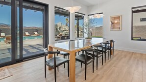 Un comedor moderno con una mesa y sillas de vidrio, ventanas amplias y vistas a una piscina y montañas.
