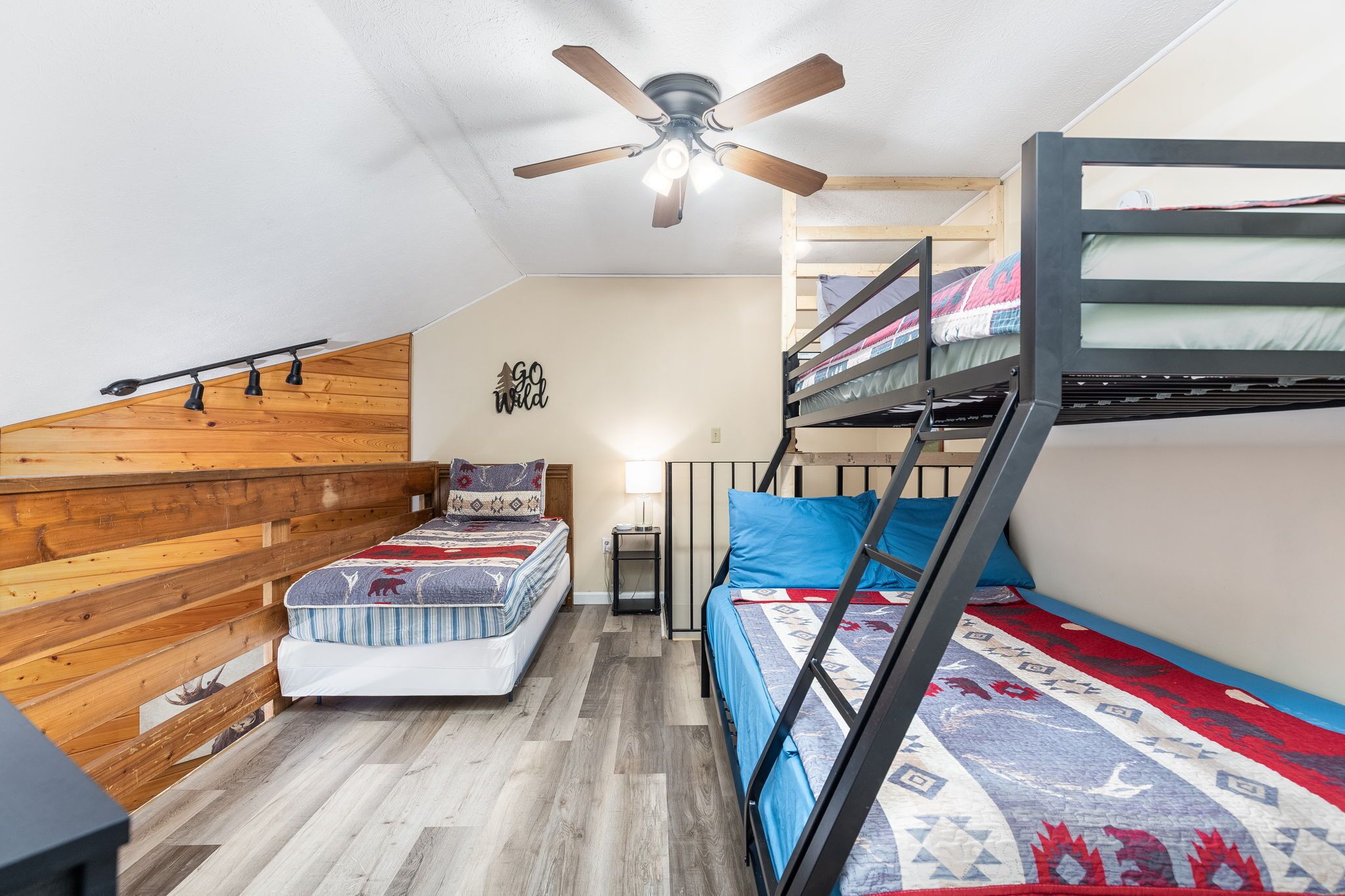 Habitación pequeña con literas, una cama individual, ventilador de techo y una mesita con lámpara.