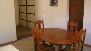 Un comedor con una mesa redonda de madera y cuatro sillas. Se observa una puerta con marco de madera y un cuadro pequeño en la pared.