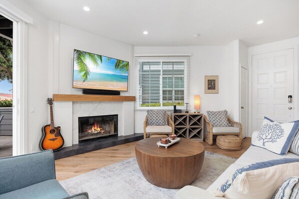 A living room with a fireplace, a television, a guitar, a round coffee table, and two seating areas with patterned cushions.