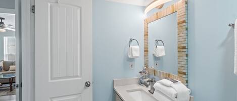 Un baño con una puerta blanca, un lavabo con espejo y toallas colgadas en ganchos.