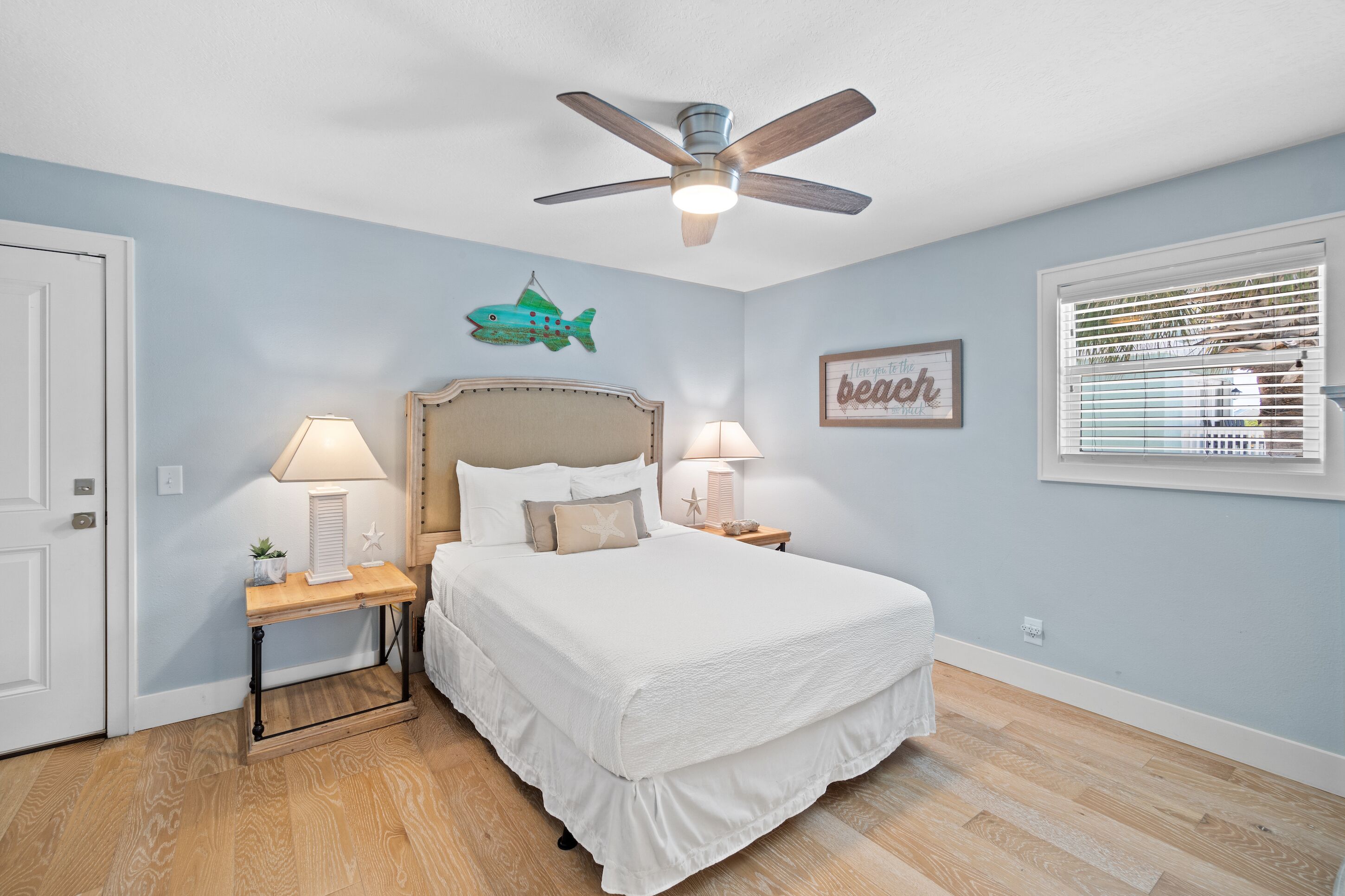 A bedroom with a bed, two bedside tables with lamps, a ceiling fan, a fish decoration, a framed quote, and a window with blinds.