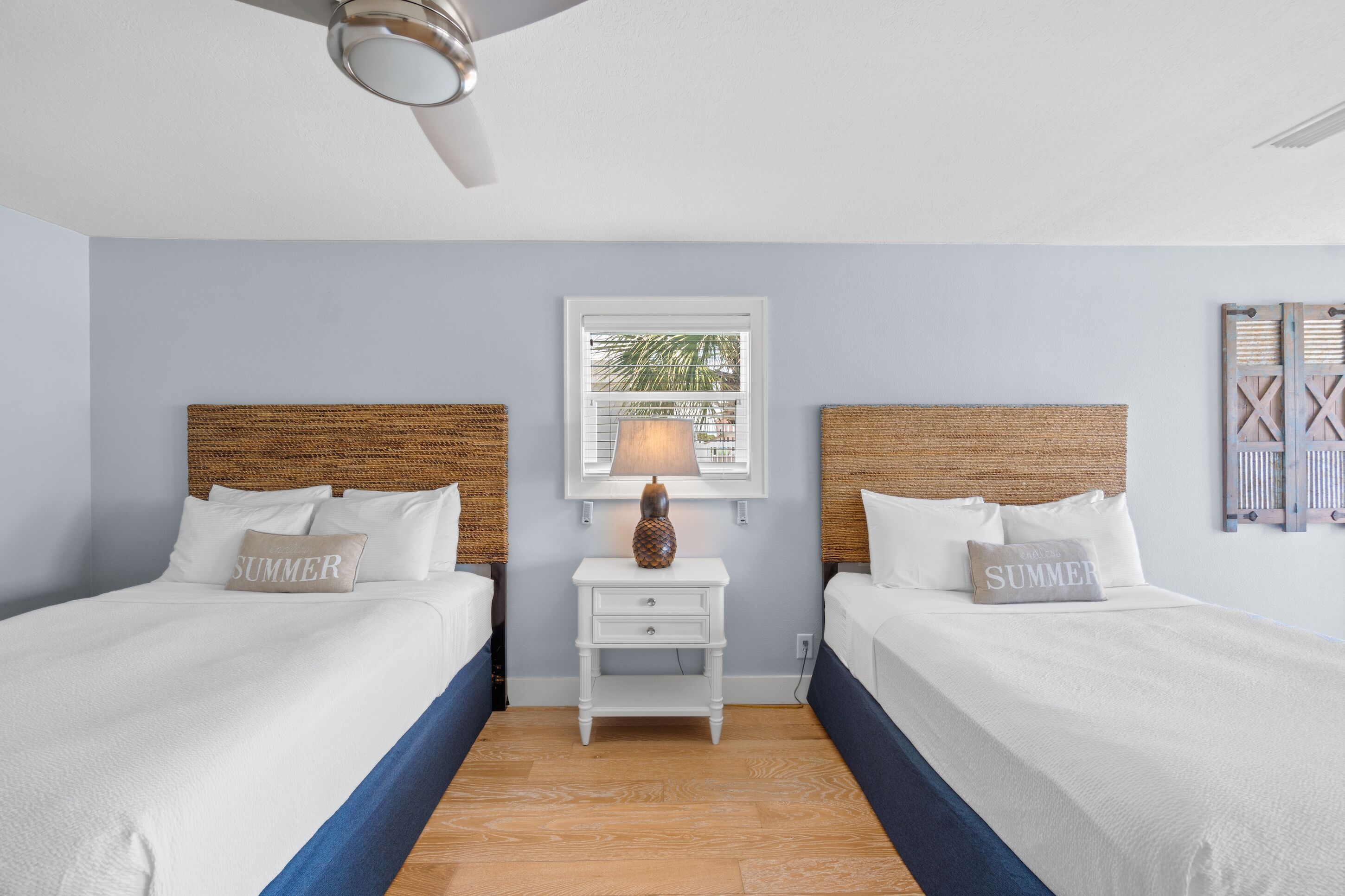 A hotel room with two beds, a small nightstand with a lamp, a framed picture on the wall, and a ceiling fan.